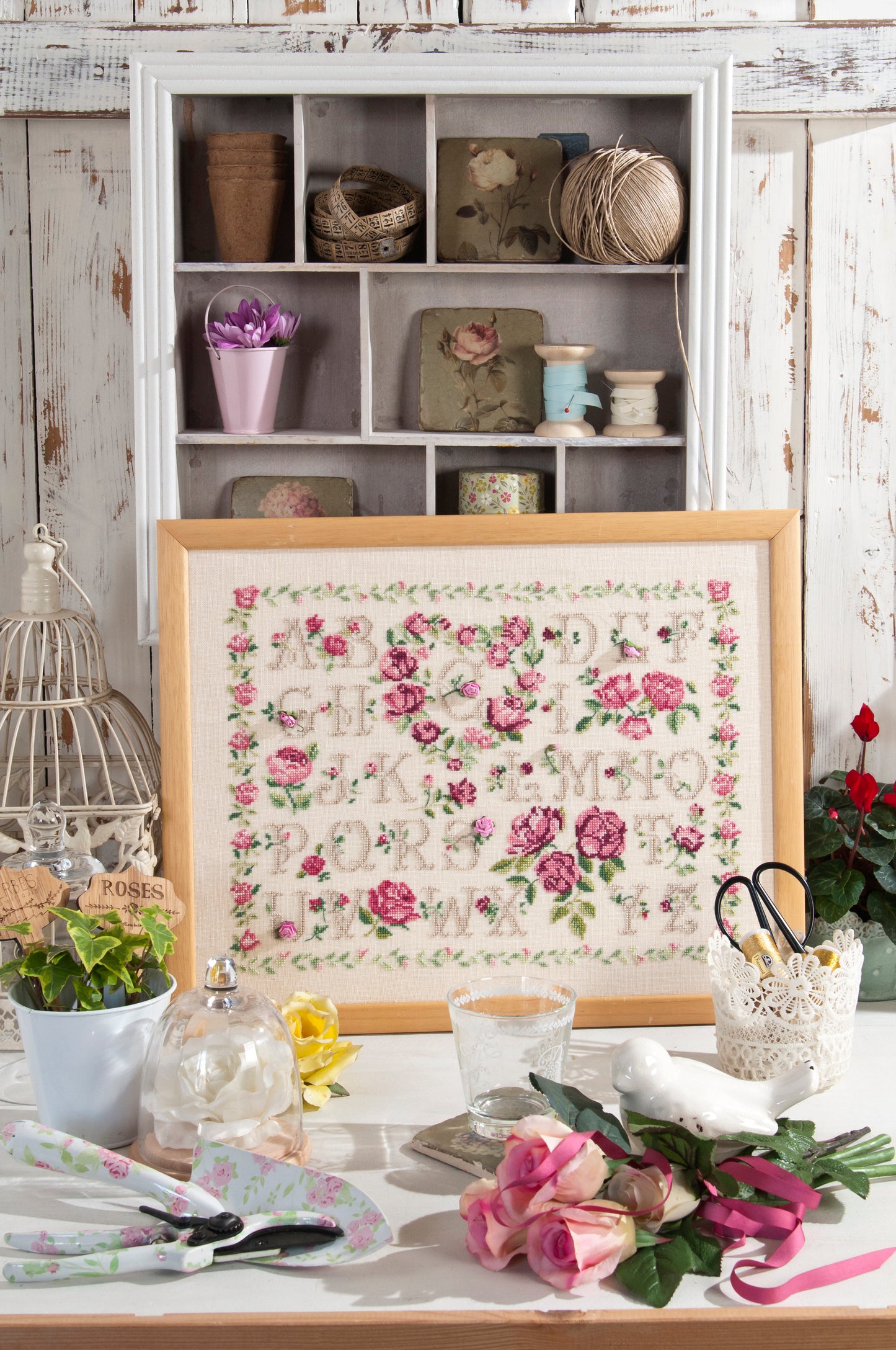 Abécédaire floral brodé sur lin naturel clair : roses anciennes, boutons et feuillages en rose tendre et vert sauge. Sampler romantique au point de croix, 40 × 45,5 cm.