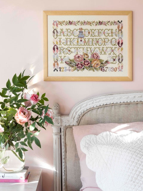 Abécédaire floral brodé sur lin écru : petite fille, roses et lettres fleuries en rose poudré et bleu tendre. Sampler romantique au point de croix, 39,1 × 30 cm.