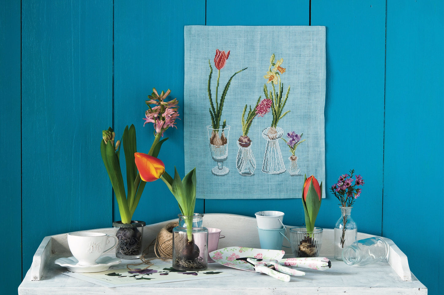 Grille point de croix – Bulbes de printemps : tulipes, narcisses, jacinthes et crocus dans des flacons en verre sur fond bleu azur – Création Point de Croix & Monique Bonnin