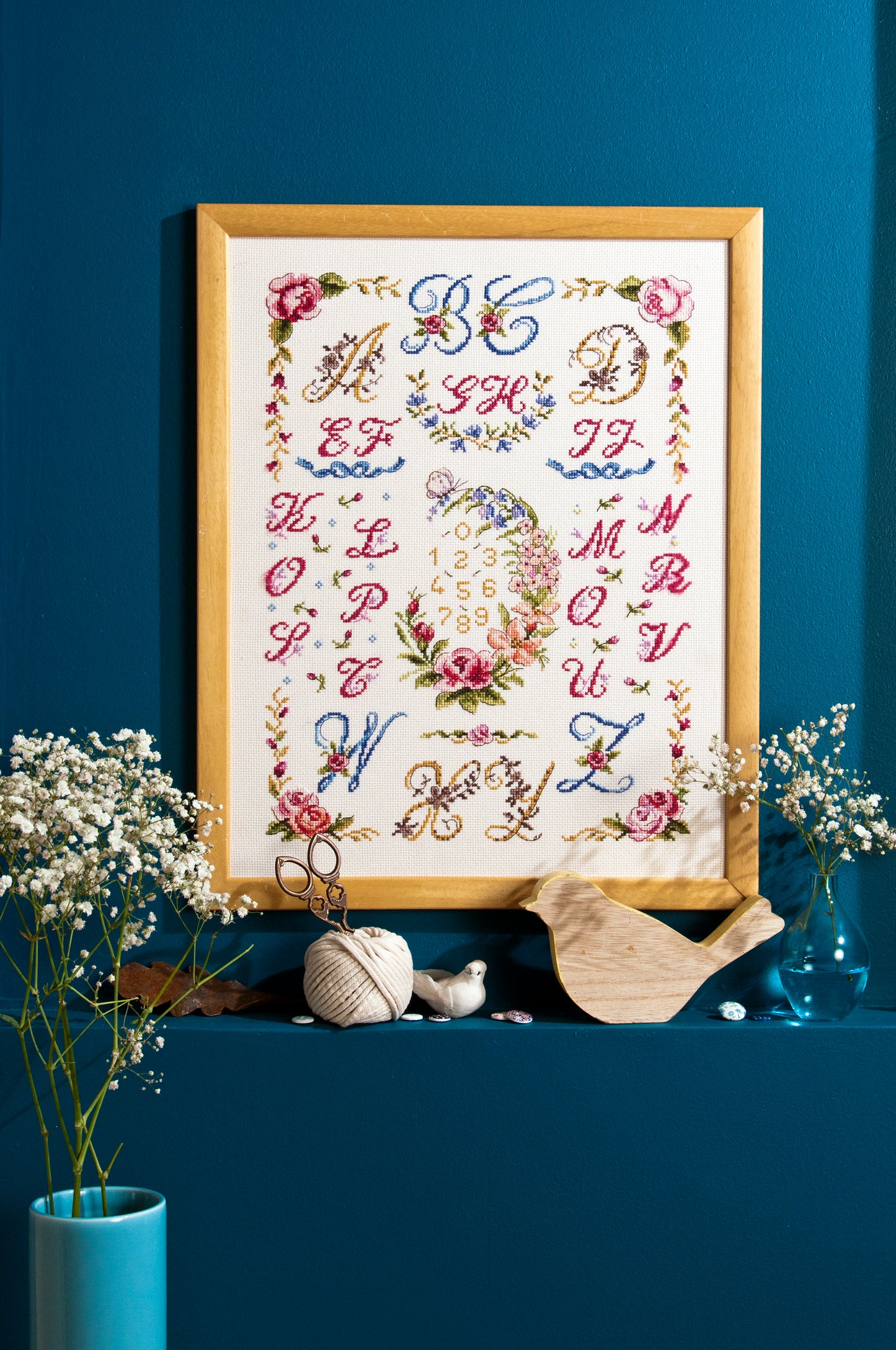 Abécédaire floral brodé sur lin clair : grandes lettres ornées et bouquets en rose poudré, bleu céleste et doré. Sampler romantique au point de croix, 32,7 × 32,7 cm.