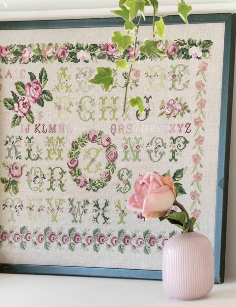 Abécédaire fleuri brodé sur lin naturel : guirlandes de roses, frises et lettres en rose poudré, mauve et vert tendre. Sampler romantique au point de croix, 38,7 × 38,7 cm.