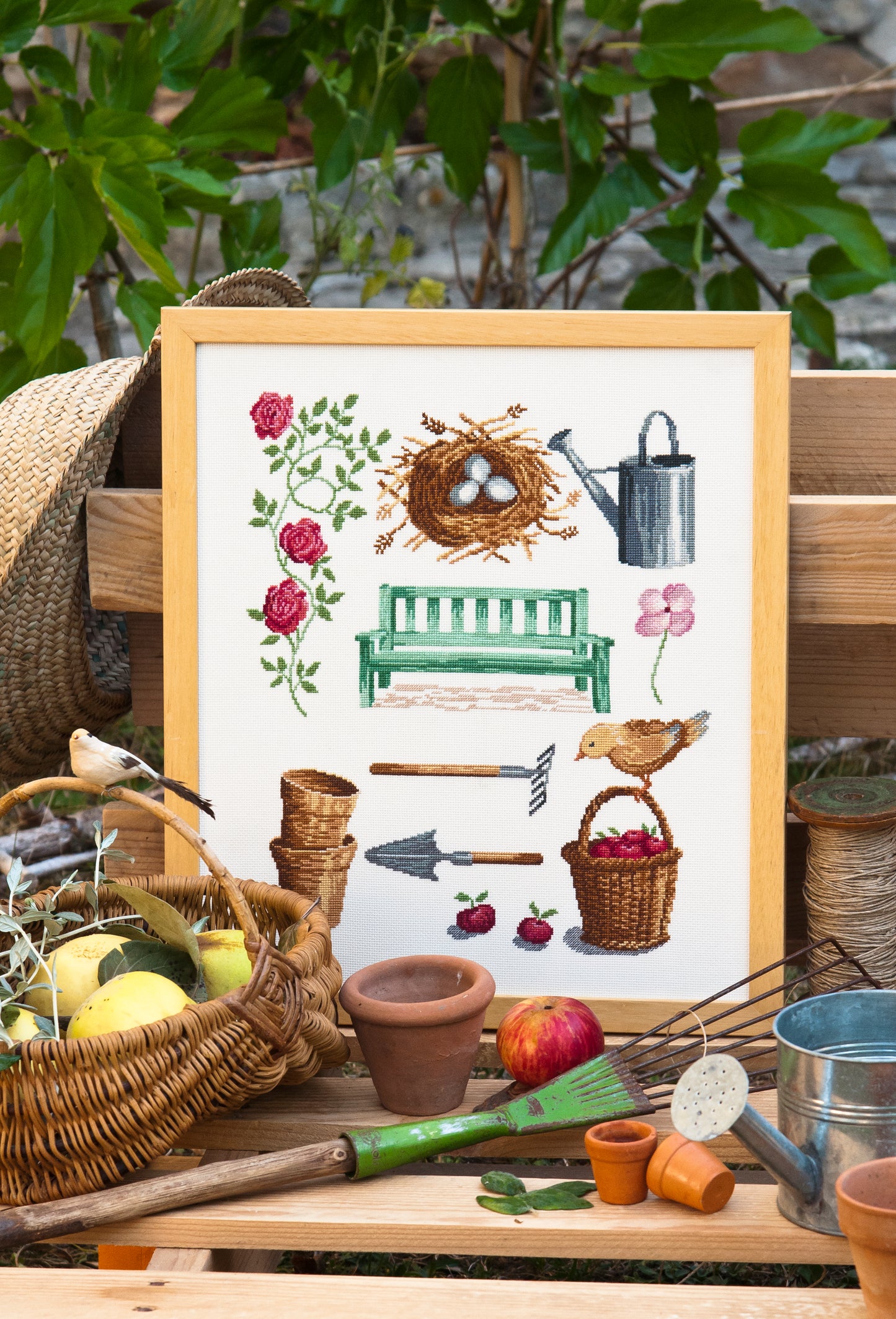 Grille point de croix – Un coin de nature : roses grimpantes, banc vert, arrosoir, panier de pommes, petit oiseau et nid avec œufs dans un jardin champêtre – Création Point de Croix