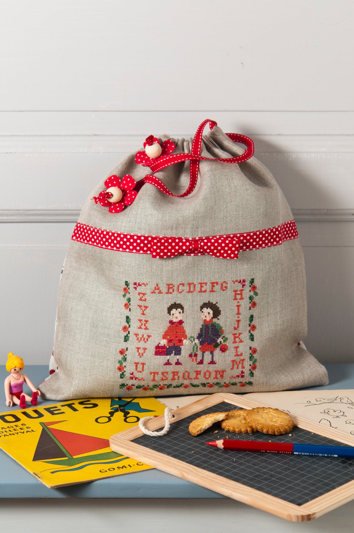 Grille de broderie point de croix : musette à goûter pour enfants, motif étiquette de cahier avec écoliers. Motif 70 x 60 pts, imprimé couleur (Réf. DMC) sur 6 pages. Illustration P. Samouiloff.