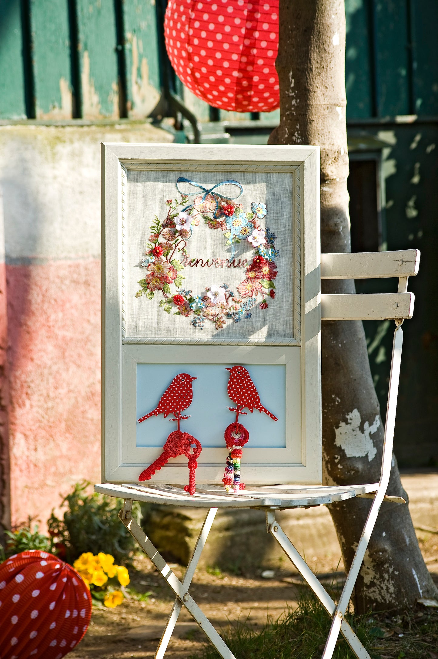 Grille broderie point de croix : tableau de bienvenue avec fleurs brodées et tissu. Motif 150x135 pts, env. 27x24 cm sur lin 11 fils ou Aïda 5,5 pts. Ref. DMC.