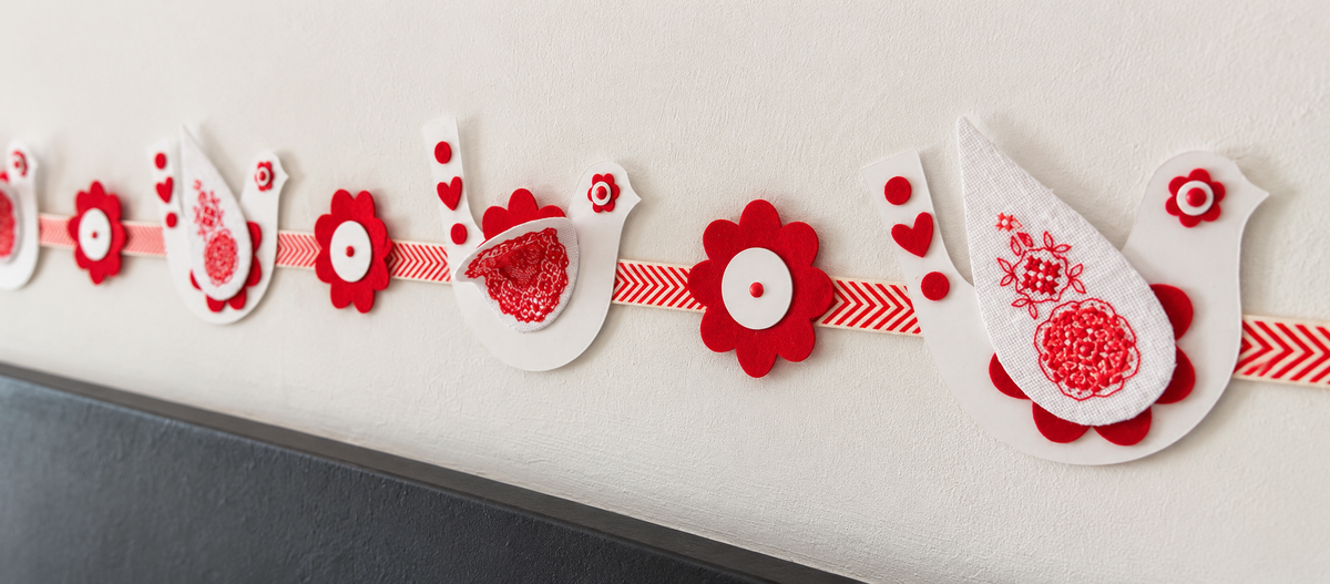 Brodez une guirlande d’oiseaux rouges et blancs d’inspiration folklorique, avec fleurs en feutrine et ruban à chevrons, pour décorer Noël.