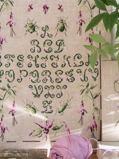 Abécédaire végétal brodé sur lin écru clair : lettres élancées, frises florales et feuillages en vert tendre et mauve. Sampler romantique au point de croix, 32,5 × 41,8 cm.