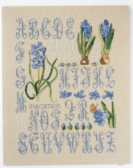 Grandes lettres ornées et jacinthes bleuées brodées sur lin naturel. Abécédaire floral romantique au point de croix, 21,8 × 32,7 cm, esprit herbier ancien.