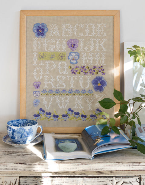 Alphabet floral brodé sur lin naturel : grandes lettres enlacées de pensées en violet et mauve. Sampler printanier romantique au point de croix, 32,7 × 32,7 cm.