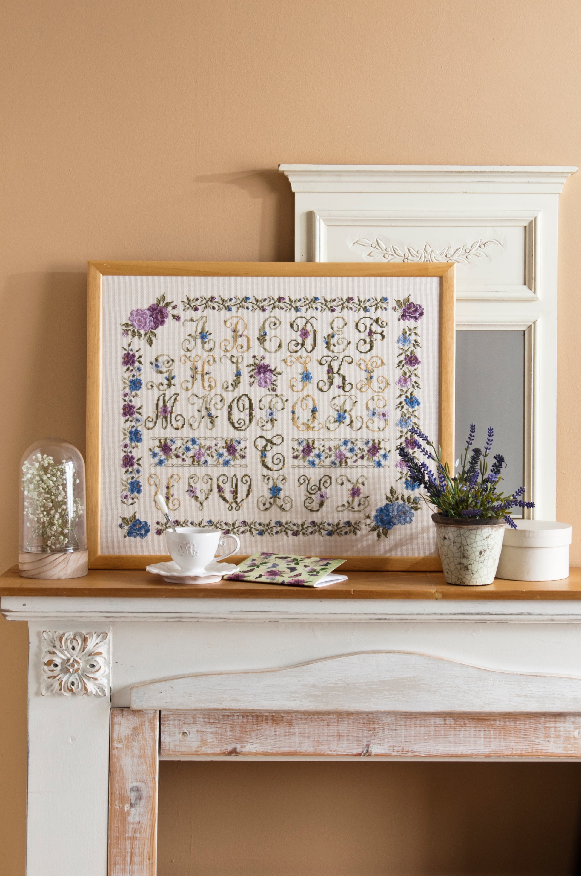 Abécédaire floral brodé sur lin clair : roses, pensées et lettres décoratives en bleu lavande et rose poudré. Sampler botanique romantique, 32,7 × 29,1 cm.