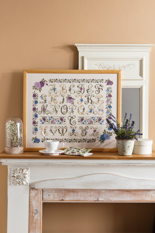 Abécédaire floral brodé sur lin clair : roses, pensées et lettres décoratives en bleu lavande et rose poudré. Sampler botanique romantique, 32,7 × 29,1 cm.