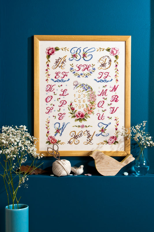 Abécédaire floral brodé sur lin clair : grandes lettres ornées et bouquets en rose poudré, bleu céleste et doré. Sampler romantique au point de croix, 32,7 × 32,7 cm.
