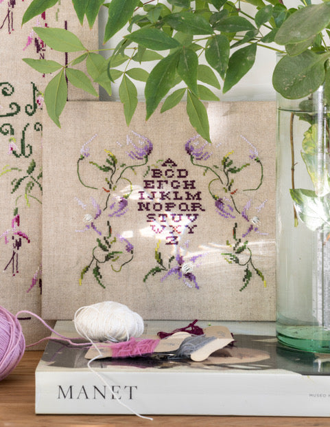 Abécédaire végétal brodé sur lin écru clair : lettres élancées, frises florales et feuillages en vert tendre et mauve. Sampler romantique au point de croix, 32,5 × 41,8 cm.