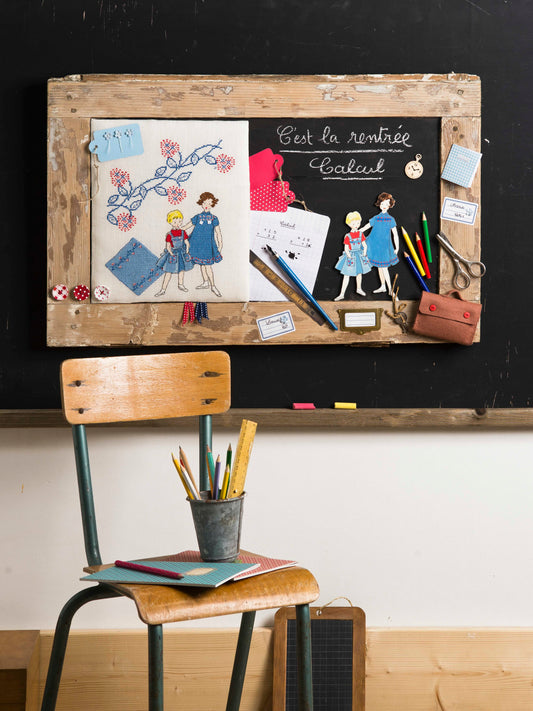 Grille point de croix – La rentrée scolaire : cartable, cahiers et plume Sergent-Major brodés sur toile de lin ivoire DMC 3865 11 fils/cm, mis en scène sur porte de meuble façon tableau noir – Création Point de Croix