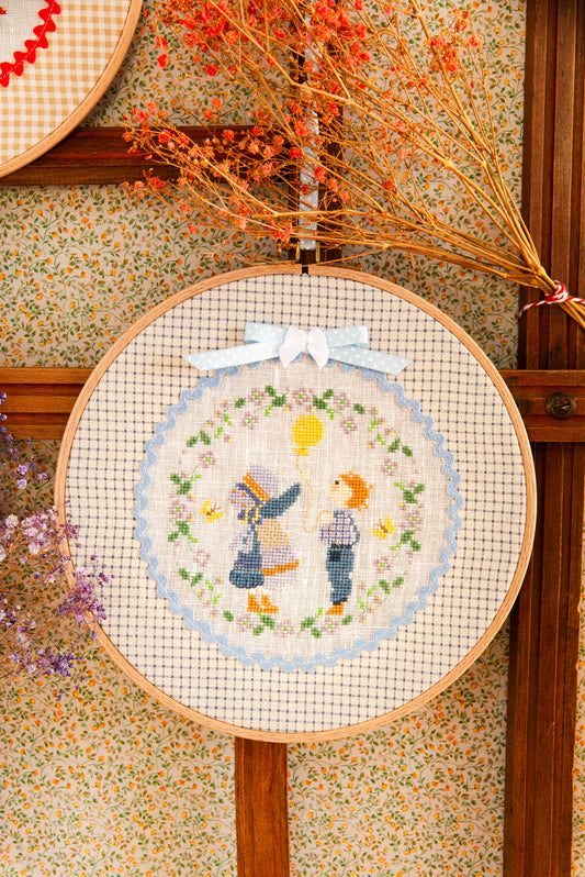 Grille point de croix : Rose et son amie au ballon dans une couronne fleurie. 74×74 pts, Aïda 5,5 pts/cm ou lin 11 fils/cm. Réf. DMC, 6 pages.