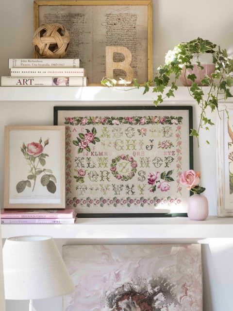 Abécédaire fleuri brodé sur lin naturel : guirlandes de roses, frises et lettres en rose poudré, mauve et vert tendre. Sampler romantique au point de croix, 38,7 × 38,7 cm.