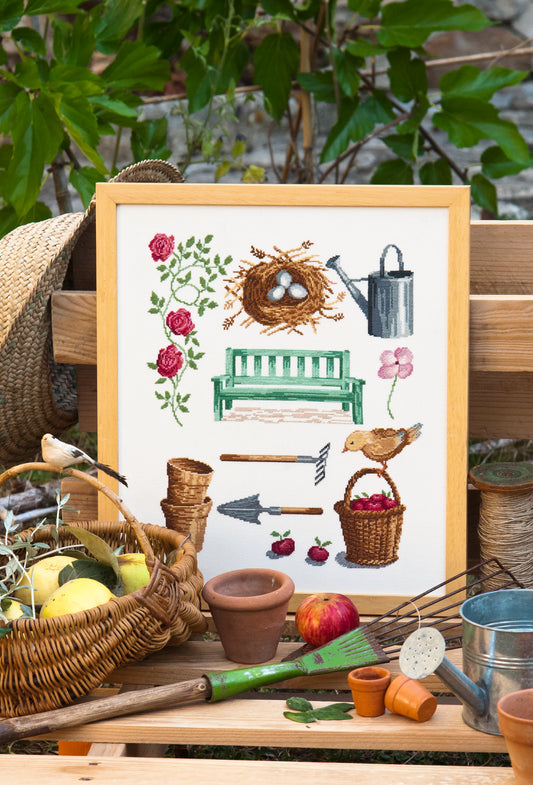 Grille point de croix – Un coin de nature : roses grimpantes, banc vert, arrosoir, panier de pommes, petit oiseau et nid avec œufs dans un jardin champêtre – Création Point de Croix