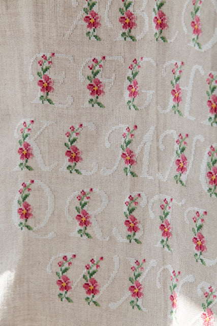 Abécédaire floral brodé sur lin naturel : lettres majuscules fleuries en rose ancien et vert feuillage. Motif romantique pour coussin, 27,3 × 27,3 cm.
