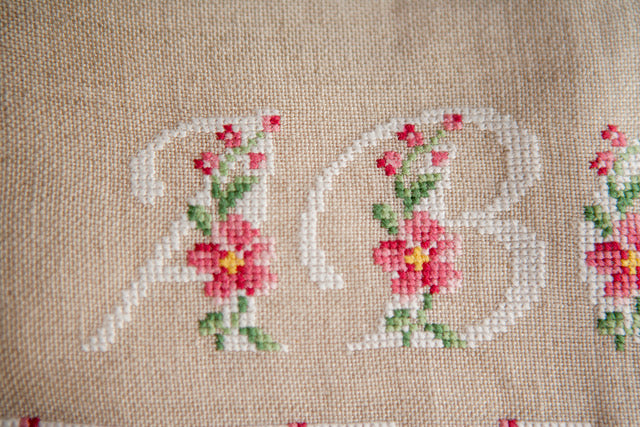 Abécédaire floral brodé sur lin naturel : lettres majuscules fleuries en rose ancien et vert feuillage. Motif romantique pour coussin, 27,3 × 27,3 cm.