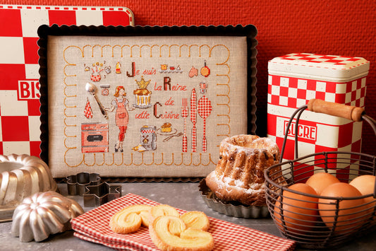 Grille broderie point de croix : tableau humoristique dans un moule à gâteau. 90 x 124 pts, imprimée en couleur sur 4 pages. Réf. DMC incluses.