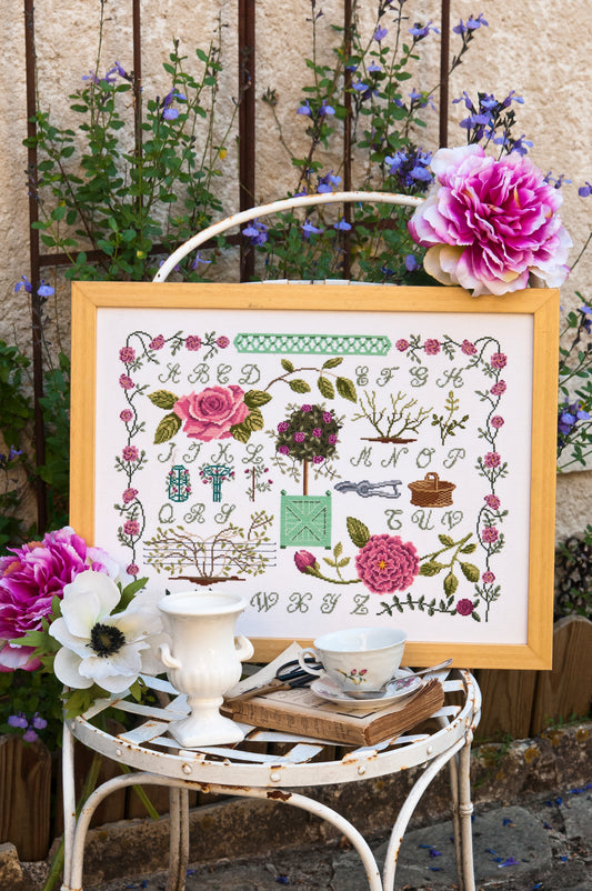 Grille point de croix – Abécédaire Roseraie : alphabet entouré de roses estivales, arbustes taillés, paniers de jardin et bordures végétales, ambiance roseraie romantique – Création Point de Croix